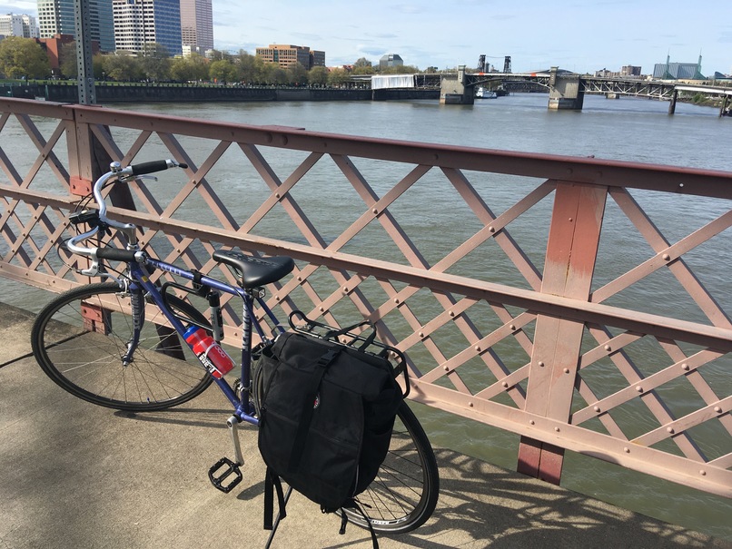 A scene of the Wilamette river from the deck of the Hawthorne Bridge looking North-West with The Morrison Bridge, Steel Bridge, The Oregon Convention center, and Big Pink clearly visible in the background. My bluish-purple Schwinn Prelued (a road bike) with oxford handle bars is pictured in the foreground against the red, weather beaten railing of the bridge.